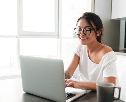 Image of happy young woman sitting at the kitchen in home. Looking aside chatting by laptop computer.